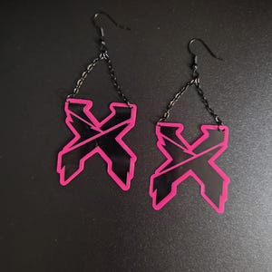 Puede incluir: Un par de pendientes colgantes con forma de letra X negra y un contorno rosa intenso. Cada pendiente cuelga de una fina cadena de metal negro sujeta a un gancho para la oreja negro, creando un diseño geométrico y llamativo para las orejas.