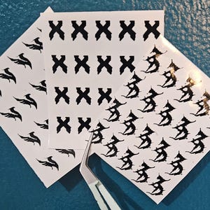 May include: Three white sheets of adhesive decals featuring black graphic designs. One sheet displays rows of black X symbols, another shows small winged icons, and the third features repeating gothic-style letter designs. A pair of metal tweezers is included.