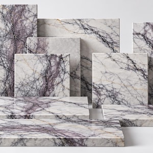 May include: A collection of rectangular and square marble slabs featuring a polished white surface with prominent, intricate purple and dark grey veining patterns. The slabs are arranged in a tiered display, showcasing various sizes and orientations.