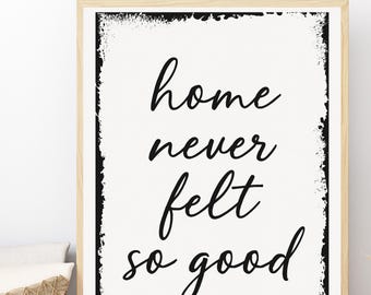 May include: A framed print with the words "home never felt so good" in black script on an off-white background. The frame is light brown, and the print has a distressed black border. The print is a decorative item.