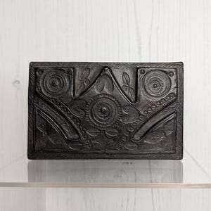 May include: A rectangular, dark-colored box with intricate, embossed floral and geometric patterns. The box features circular and curved designs, with a textured surface. The box is displayed on a clear, acrylic surface against a white wooden backdrop.