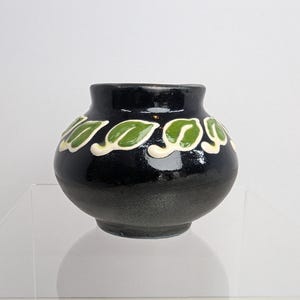 Peut inclure: Un petit vase en céramique noir, de forme arrondie et doté d'une finition brillante. La partie supérieure du vase présente une bande décorative répétitive composée de feuilles en relief peintes en vert et soulignées de blanc.