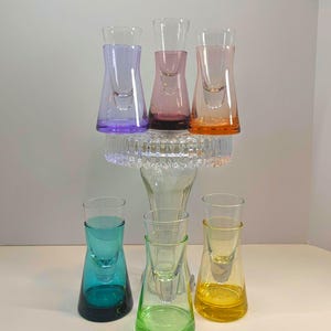 May include: A collection of colorful shot glasses in shades of purple, pink, orange, teal, green, and yellow. The glasses are arranged on a clear glass stand, showcasing their vibrant hues and unique design. Ideal for a home bar or party.