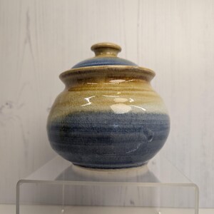 May include: A handcrafted ceramic jar with a lid. The jar is round with a blue base, a tan middle, and a blue lid. The jar is sitting on a clear acrylic shelf. The jar is likely used for storage.