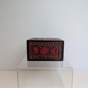 May include: A rectangular wooden box featuring a dark black frame and a central panel with a deep red background. The surface is decorated with intricate black carved patterns, including a central diamond shape flanked by heart-like motifs.