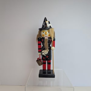 May include: A wooden nutcracker figure dressed as a witch, featuring a black pointed hat with a white cat, a black coat with red stars, red and black striped sleeves and legs, a long brown beard, a broom, and a small bucket with red items.