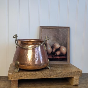 May include: A hammered copper pot with a rounded body, a wide opening, and a brass swing handle. The pot features three curved brass feet attached to the base. The exterior surface has a polished, reflective finish with visible metal texture.