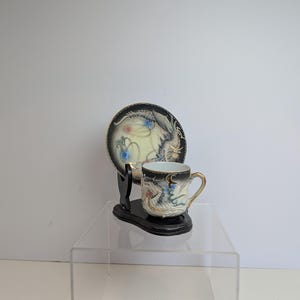 May include: A porcelain teacup and matching saucer set featuring a raised relief dragon design in white, gold, and blue against a black background. The set is displayed on a black wooden stand, showcasing intricate detailing on the cup and saucer.