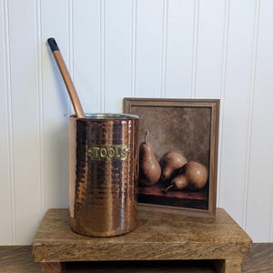 May include: A cylindrical copper container with a hammered texture and a brass plaque embossed with the word TOOLS. A long, thin copper-colored utensil with a black handle rests inside. A small framed painting of three pears sits nearby.