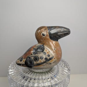 May include: A ceramic bird figurine with a brown body, black beak, and decorative wing patterns in black and orange. The bird is perched on a clear, glass pedestal.