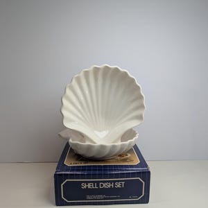 May include: A two-piece white ceramic shell dish set featuring a scalloped edge design. The dishes are displayed resting on a blue rectangular cardboard box with white grid lines and the text SHELL DISH SET printed on the front panel.