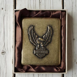 May include: A rectangular, brass-colored metal box with a raised Harley Davidson eagle emblem. The box is nestled in a satin-like brown fabric within a white cardboard box, set against a rustic wooden background.
