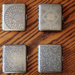 May include: Four rectangular metal cases with hinged lids, each featuring a distinct engraved design. One displays a compass rose, another a floral scroll pattern, one a circular religious emblem, and the last an intricate leaf motif.