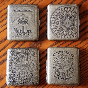 May include: Four antique brass cigarette cases. One case features the Marlboro Lights logo and text. Another has a compass design with the text "Sailing to the World". The third case has a circular design with text. The last case has a Marine Corps emblem.