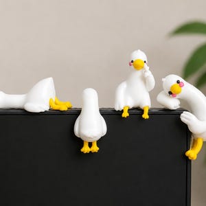 Puede incluir: Cuatro pequeñas figuras de pato blancas con picos y patas amarillas, y manchas rosas en las mejillas. Un pato está acostado, uno mira hacia otro lado, uno está sentado con un ala cerca de la cara y uno se inclina sobre el borde negro.