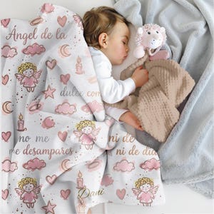 May include: A sleeping baby is nestled under a white blanket with pink angel and heart patterns. The blanket has text in Spanish. A beige plush toy and a light blue blanket are also visible.