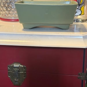 May include: A sage green rectangular planter with three small feet. The planter has a simple, geometric design. The planter is sitting on a white surface. A red trunk with a metal clasp is in the background.