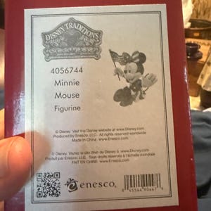 Puede incluir: Caja roja con una etiqueta blanca que presenta una figura de Minnie Mouse de Disney Traditions. La figura sostiene una peque&ntilde;a bandera estadounidense. La etiqueta incluye el texto "Minnie Mouse Figurine" y el logotipo de Enesco.