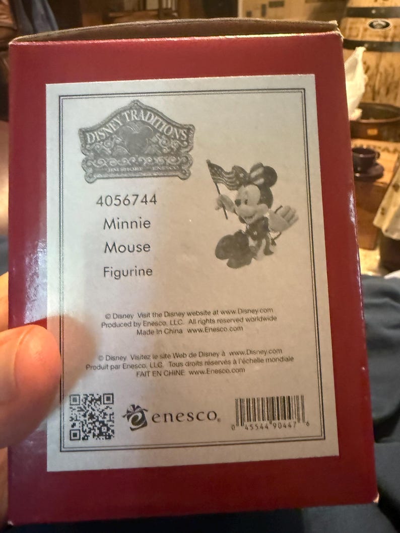Puede incluir: Caja roja con una etiqueta blanca que presenta una figura de Minnie Mouse de Disney Traditions. La figura sostiene una peque&ntilde;a bandera estadounidense. La etiqueta incluye el texto "Minnie Mouse Figurine" y el logotipo de Enesco.