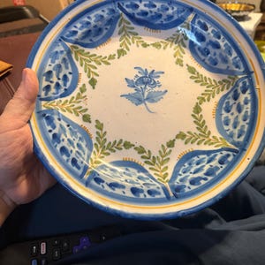 May include: A round, hand-painted ceramic plate with a white center and a blue and yellow rim. The plate features a floral design in blue and green, with a central flower and surrounding leaf patterns. The plate is held by a hand.
