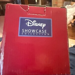 Puede incluir: Una caja rectangular roja con el logotipo de Disney Showcase Collection en un rect&aacute;ngulo azul. La caja tiene un peque&ntilde;o desgarro en la esquina superior izquierda. El fondo est&aacute; borroso.