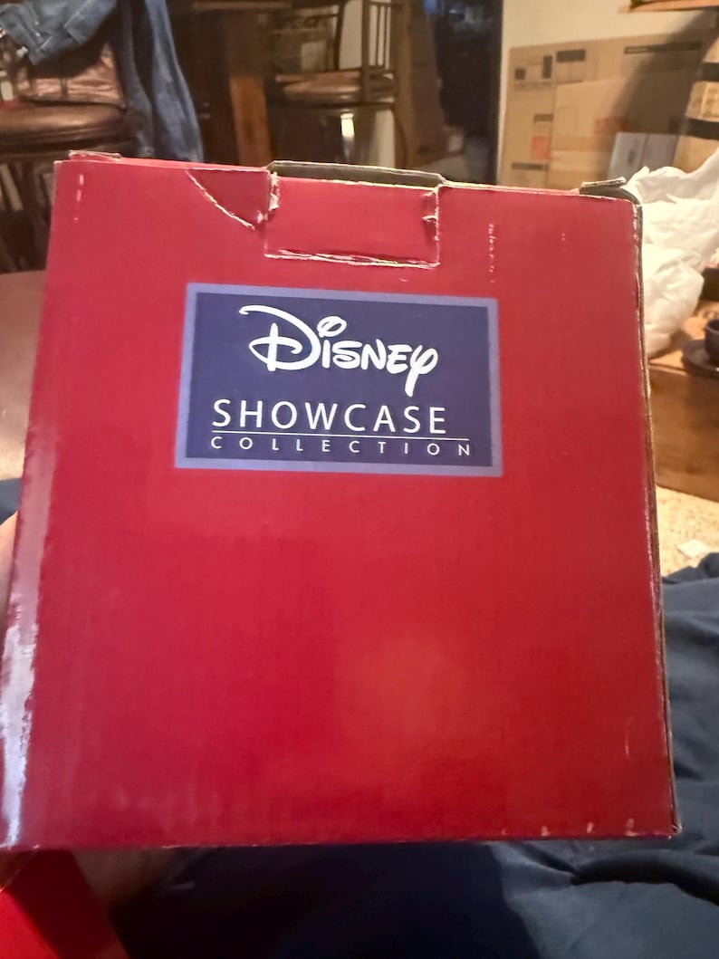 Puede incluir: Una caja rectangular roja con el logotipo de Disney Showcase Collection en un rect&aacute;ngulo azul. La caja tiene un peque&ntilde;o desgarro en la esquina superior izquierda. El fondo est&aacute; borroso.
