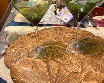 May include: Two green martini glasses with clear stems sit on a wooden table carved with lily pads and a frog. The glasses have a classic triangular bowl shape. The table has a natural wood finish.