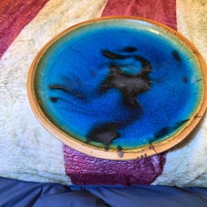 May include: A round ceramic plate with a vibrant blue center and a dark, abstract design. The plate has a light brown rim and a glossy finish. The plate is a decorative item.