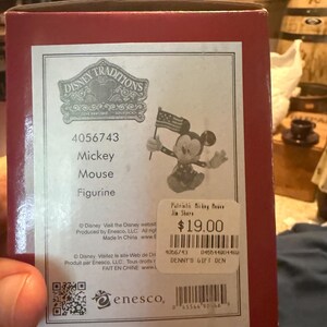 Puede incluir: Una caja roja con una figura de Disney Traditions. La etiqueta de la caja muestra una figura de Mickey Mouse sosteniendo una peque&ntilde;a bandera estadounidense. El texto en la caja dice "Mickey Mouse Figurine" y "Patriotic Mickey Mouse Jin Shore."