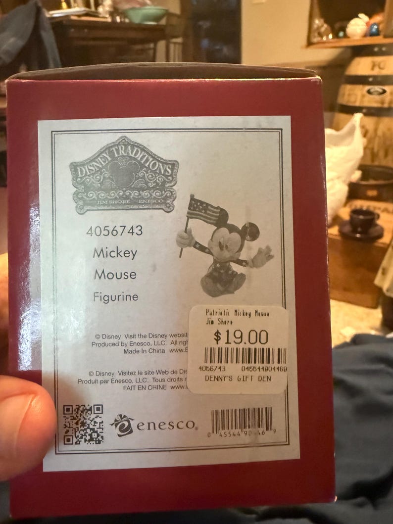 Puede incluir: Una caja roja con una figura de Disney Traditions. La etiqueta de la caja muestra una figura de Mickey Mouse sosteniendo una peque&ntilde;a bandera estadounidense. El texto en la caja dice "Mickey Mouse Figurine" y "Patriotic Mickey Mouse Jin Shore."
