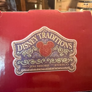 Puede incluir: Una caja roja con una etiqueta decorativa con las palabras "Disney Traditions" en una fuente estilizada. La etiqueta incluye una cabeza de Mickey Mouse y el texto "designed by Jim Shore for Enesco."