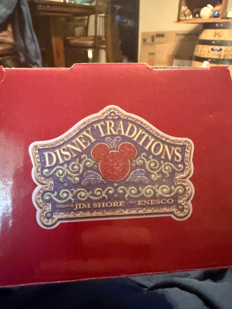Puede incluir: Una caja roja con una etiqueta decorativa con las palabras "Disney Traditions" en una fuente estilizada. La etiqueta incluye una cabeza de Mickey Mouse y el texto "designed by Jim Shore for Enesco."
