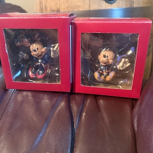 Puede incluir: Dos cajas rojas que contienen cada una una figura de Mickey Mouse. Las figuras visten atuendos patri&oacute;ticos y sostienen banderas americanas. Las cajas est&aacute;n sobre una superficie de cuero marr&oacute;n, con un barril de madera al fondo.