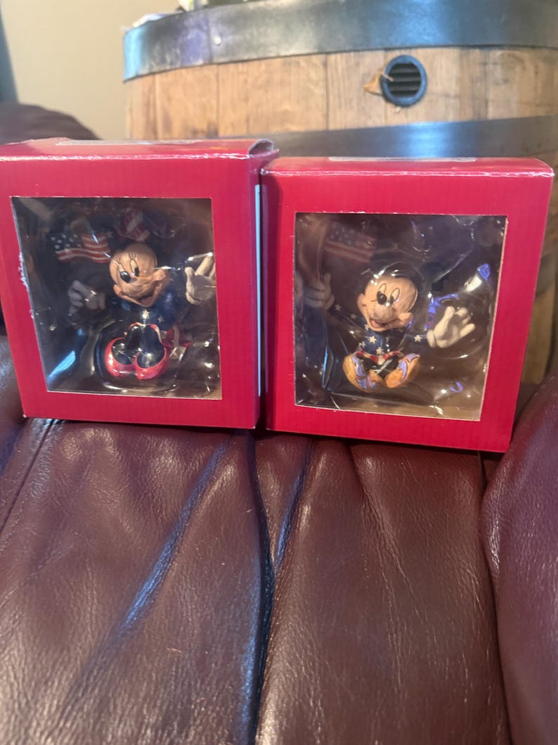 Puede incluir: Dos cajas rojas que contienen cada una una figura de Mickey Mouse. Las figuras visten atuendos patri&oacute;ticos y sostienen banderas americanas. Las cajas est&aacute;n sobre una superficie de cuero marr&oacute;n, con un barril de madera al fondo.