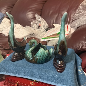 May include: Three ceramic swan figurines in shades of teal and brown. The swans have a glossy finish and are arranged on a blue towel. The swans are on a wooden box with gold hardware.