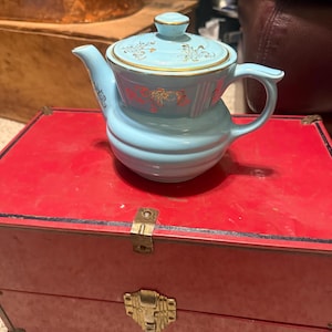 May include: A light blue ceramic teapot with a lid, featuring gold floral designs. The teapot sits on a red box with gold hardware. The teapot has a handle and a spout.