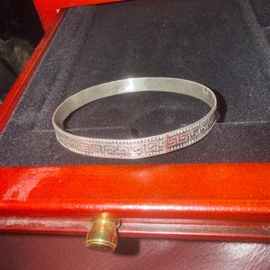May include: A silver bangle bracelet with a Greek key design. The bracelet is displayed on a black velvet surface inside a red wooden jewelry box. The bracelet's design is a repeating pattern of interlocking squares and lines.