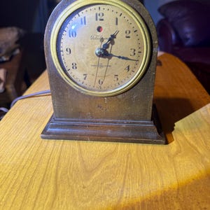 May include: An antique Telechron clock with a wooden frame and a gold-colored clock face. The clock face has black numerals and hands, with the Telechron logo in the center. The clock is sitting on a wooden table.