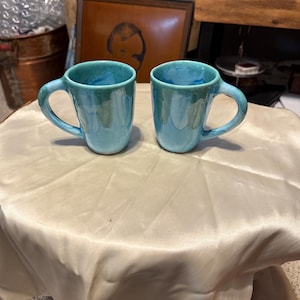 May include: Two ceramic mugs with a turquoise glaze. Each mug has a rounded body, a curved handle, and a glossy finish. The mugs are displayed on a cream-colored satin surface.