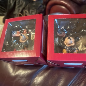 Puede incluir: Dos cajas rojas con ventanas transparentes que muestran figuras de Mickey Mouse. Cada figura viste ropa patri&oacute;tica y sostiene una bandera estadounidense. Las figuras est&aacute;n sobre un fondo oscuro.
