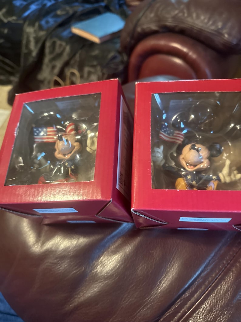 Puede incluir: Dos cajas rojas con ventanas transparentes que muestran figuras de Mickey Mouse. Cada figura viste ropa patri&oacute;tica y sostiene una bandera estadounidense. Las figuras est&aacute;n sobre un fondo oscuro.