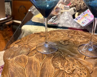 May include: Two blue-tinted martini glasses with long stems sit on a wooden tray. The tray is carved with a leaf and frog design. The glasses have a classic triangular bowl shape.