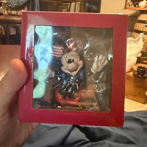 Puede incluir: Una figura de Minnie Mouse en una caja roja. La figura lleva un atuendo patri&oacute;tico con una combinaci&oacute;n de colores rojo, blanco y azul. La caja es roja y tiene una ventana de pl&aacute;stico transparente.