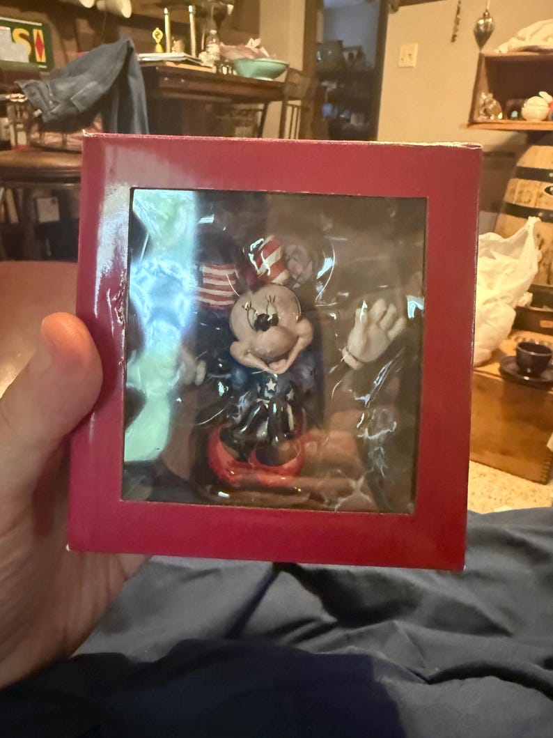 Puede incluir: Una figura de Minnie Mouse en una caja roja. La figura lleva un atuendo patri&oacute;tico con una combinaci&oacute;n de colores rojo, blanco y azul. La caja es roja y tiene una ventana de pl&aacute;stico transparente.
