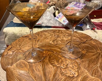 May include: Two amber-colored martini glasses with long stems sit on a brown, carved wooden table. The table features a lotus leaf design with a frog sculpture. The glasses are empty.