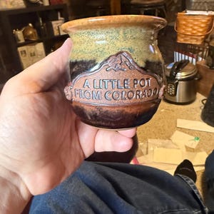May include: A handmade ceramic pot with a mountain design and the text "A Little Pot From Colorado." The pot has a brown, black, and tan color scheme. The pot is held in a person's hand.