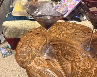 May include: A martini glass with a purple-tinted bowl and stem, filled with ice and a dark liquid. The glass sits on a round, wooden table with carved lotus leaves and a frog design. The table has a dark brown finish.