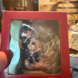 Puede incluir: Una caja roja que contiene una figura de Mickey Mouse. Mickey viste un atuendo patri&oacute;tico con una chaqueta azul con estrellas blancas y sostiene una peque&ntilde;a bandera estadounidense. La figura est&aacute; encerrada en pl&aacute;stico transparente.