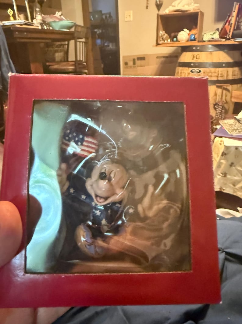 Puede incluir: Una caja roja que contiene una figura de Mickey Mouse. Mickey viste un atuendo patri&oacute;tico con una chaqueta azul con estrellas blancas y sostiene una peque&ntilde;a bandera estadounidense. La figura est&aacute; encerrada en pl&aacute;stico transparente.