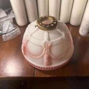 May include: A dome-shaped milk glass lamp shade featuring a soft pink gradient and raised white relief patterns of swags and tassels. The top includes a circular brass-toned metal fitter with small screws for attachment to a lamp fixture.